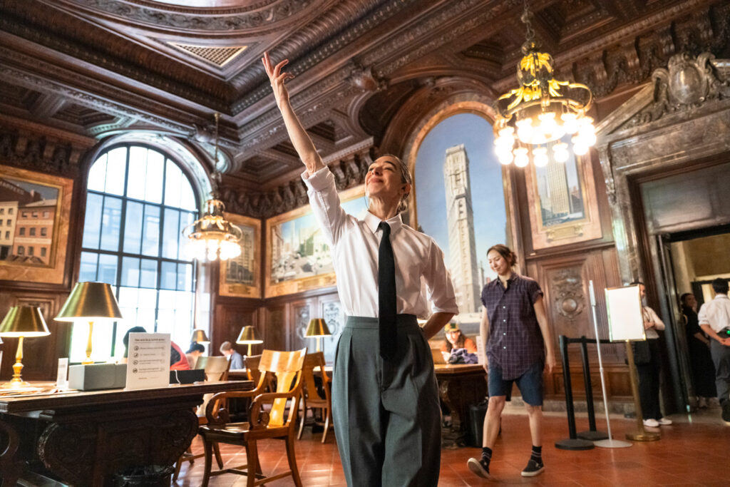 Barnes, wearing a button-down shirt and tie, stands in an ornate library room. She extends her right hand toward the ceiling, a small smile on her face.