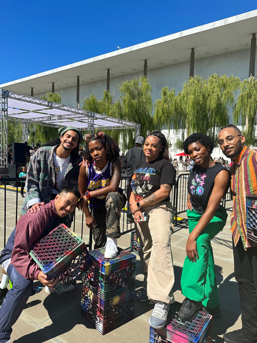 A group of dancers in casual clothes poses in front of the Kennedy Center, some holding or resting their feet atop several multicolored plastic crates.